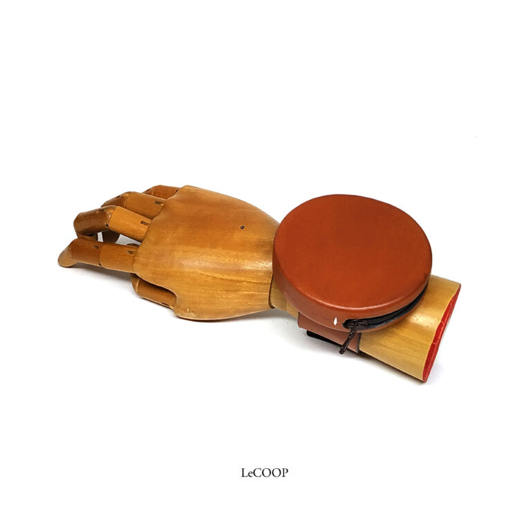 Monedero redondo de cuero color cuero con cremallera, sobre una mano articulada de madera