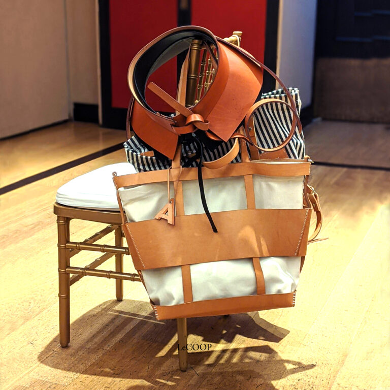 Bolsos de lona y cuero color cuero con una inicial colgada y cinturones, apilados sobre una silla