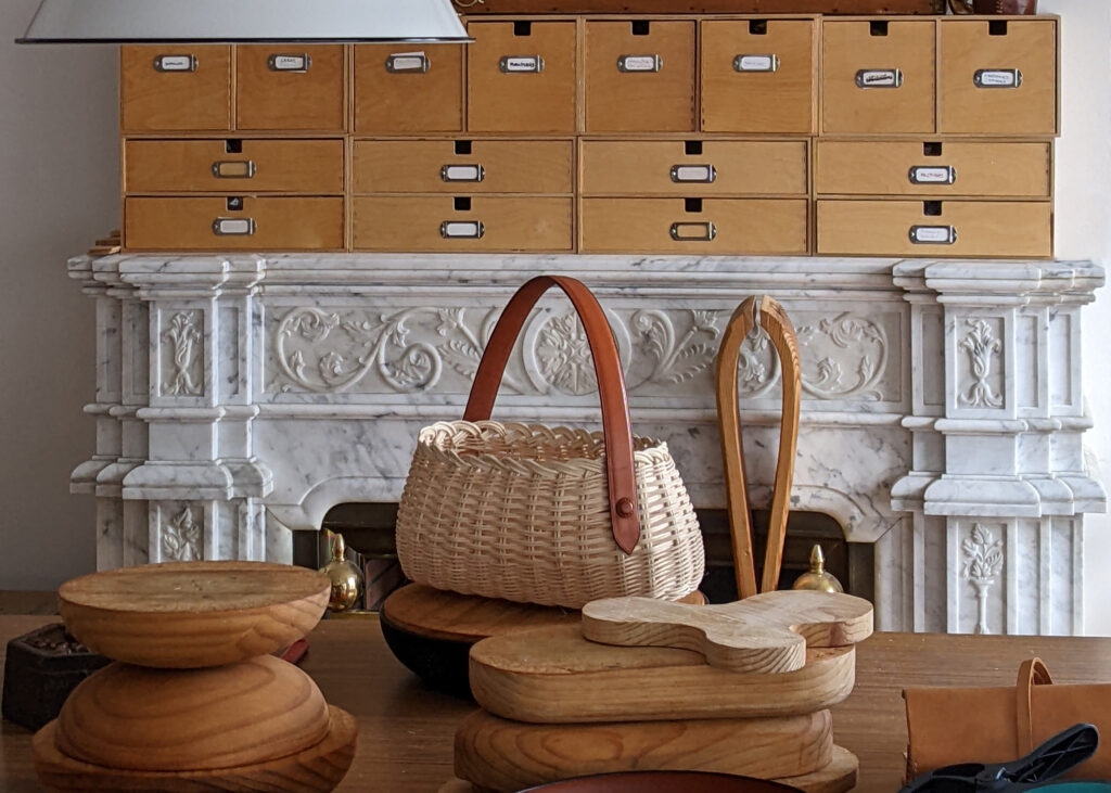 Bolso cesta con asa de cuero y hormas de madera delante de la chimenea de mármol del taller